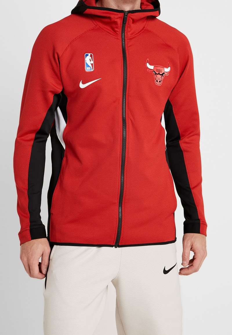 Homme portant un sweat à capuche zippé rouge des Chicago Bulls avec des accents noirs et blancs et un pantalon Nike blanc avec un logo swoosh noir.