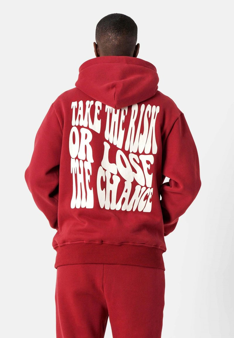 Sudadera roja con texto en negrita blanco "TOMA EL RIESGO O PIERDE LA OPORTUNIDAD" en la espalda. Material de algodón, corte relajado, bolsillo canguro, puños acanalados.