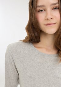 Giovane donna con pelle chiara e capelli castani, che indossa una maglietta grigia chiara a girocollo, rivolta verso la fotocamera con un'espressione neutra.