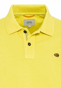 Gelbes Poloshirt aus strukturiertem Stoff, mit einer Knopfleiste mit drei Knöpfen und einem kleinen bestickten Logo auf der linken Brust.