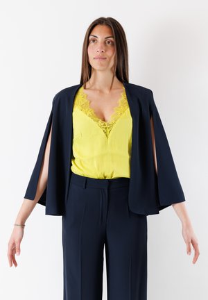 Giacca aperta blu navy con maniche larghe, sovrapposta a un top giallo con bordi in pizzo. Pantaloni blu navy su misura completano l'outfit.