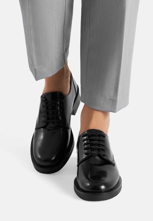 Schwarze Schnürschuhe aus Leder mit glänzender Oberfläche, rund zulaufender Spitze und robuster Gummisohle. Kombiniert mit hellgrauen, maßgeschneiderten Hosen.