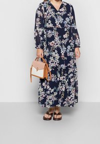 Námořnické květinové maxi šaty s dlouhými rukávy a výstřihem do V; spárované s béžovou a hnědou kabelkou; modelka má na sobě hnědé sandály.