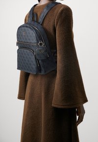 Zaino in tessuto blu navy con logo in rilievo, due tasche frontali con cerniera, un portachiavi rotondo e spallacci imbottiti e regolabili, indossato su un indumento marrone.