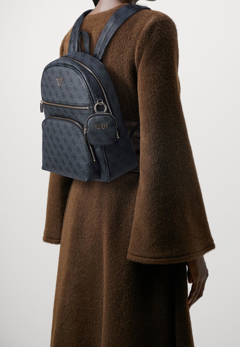 Zaino in tessuto blu navy con logo in rilievo, due tasche frontali con cerniera, un portachiavi rotondo e spallacci imbottiti e regolabili, indossato su un indumento marrone.