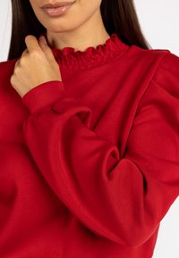 Rotes Langarmoberteil mit gefälteltem Ausschnitt, gerafften Bündchen und glatter Stofftextur. Nahaufnahme hebt die Designdetails hervor.
