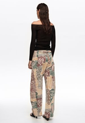 Ženska z dolgimi lasmi nosi črno bluzo brez ramen in široke hlače z visokim pasom ter večbarvnim abstraktnim čipkastim vzorcem, stoji obrnjena stran.