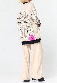 Cardigan beige avec bordure noire présentant un motif de chat noir et un détail central de chat rose, porté avec un pantalon large crème.