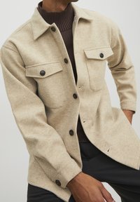 Beige wool flannel shirt jacket with two chest pockets, black buttons, and a slightly textured finish. Features a relaxed fit and fold-down collar.