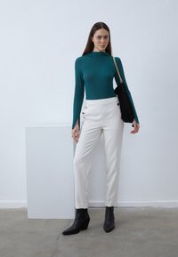 Long-sleeve teal top, white tailored pants with gold accents, and black heeled ankle boots. Background is a plain white wall.