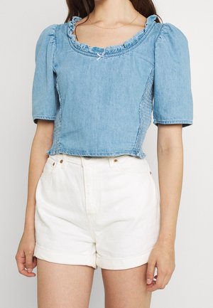 Femme portant un haut en denim bleu clair à manches bouffantes avec un col à volants et un short taille haute blanc avec ourlets roulés.