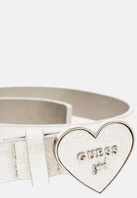 Cintura in finta pelle bianca con fibbia a forma di cuore argentata lucida, incisa con "GUESS girl." Il materiale ha una finitura texture sottile.