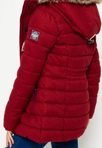 Czerwona kurtka puchowa z kapturkiem z sztucznego futra, pikowanym wzorem, kieszeniami na suwak i naszywką z logo na rękawie. Materiał o fakturze dającej ciepłe wrażenie.