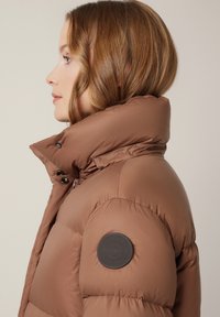 Brązowa kurtka puchowa z wysokim kołnierzem, z naszywką z logo na rękawie i błyszczącą, pikowaną fakturą. Zamek błyskawiczny i zapinanie na napy.