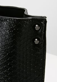 Coin d'un sac en similicuir noir avec un motif gaufré en forme d'écailles et deux rivets noirs brillants sur un fond clair.