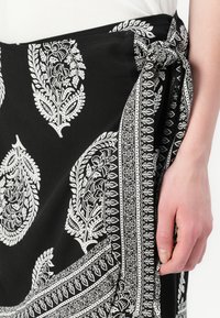 Svart kjol av tyg med intrikata vita paisley- och blommönster, knuten i midjan med en tygknut. Slät textur.