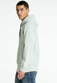 Sudadera verde claro con capucha ajustable, puños acanalados y costuras laterales. Usada sobre una camiseta blanca, combinada con jeans azules.