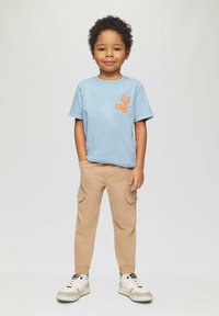 Jong kind dat met één hand in de zak staat, een lichtblauw T-shirt, beige cargobroek en witte sneakers draagt op een effen achtergrond.