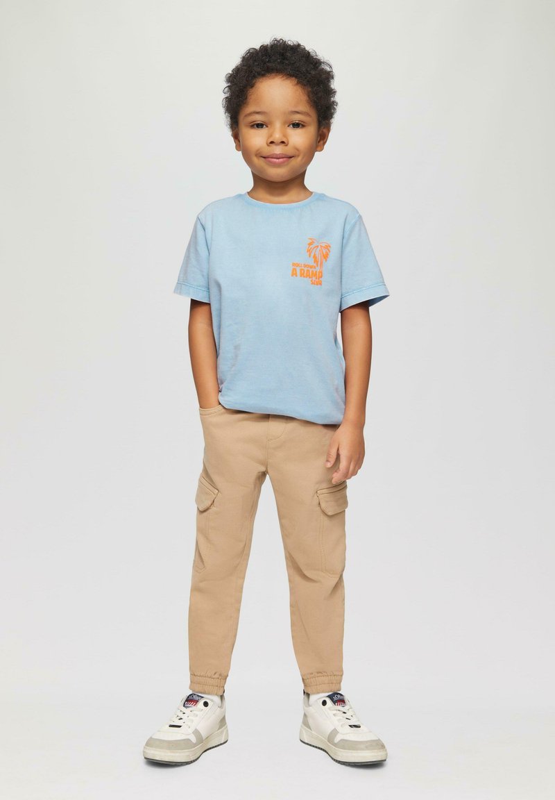 Jong kind dat met één hand in de zak staat, een lichtblauw T-shirt, beige cargobroek en witte sneakers draagt op een effen achtergrond.