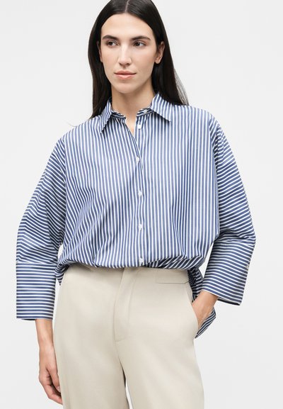Γυναίκα με μακριά σκούρα μαλλιά που φοράει μπλε και λευκό κάθετα ριγωτό πουκάμισο με κουμπιά και μπεζ παντελόνια, με το ένα χέρι στην τσέπη.
