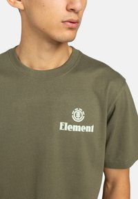 Olivovo zelené bavlnené tričko s krátkymi rukávmi, s bielym logom Element a grafikou stromu na ľavej strane hrude.