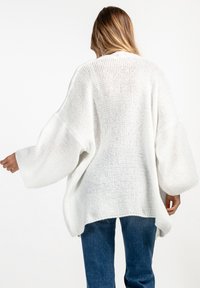 Biały sweter dzianinowy o oversize'owym kroju, z szerokimi rękawami, teksturowanym wzorem i otwartym tyłem, w połączeniu z niebieskimi dżinsami.