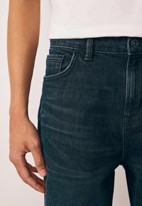 Jeans din denim închis, cu o croială slim, ușoară uzură și un design clasic cu cinci buzunare, având detalii vizibile ale butonului și taliei.