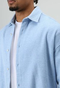 Lichtblauwe overhemdjas van een wolmix met een puntkraag, drukknopen en een zachte textuur; gedragen over een wit T-shirt.