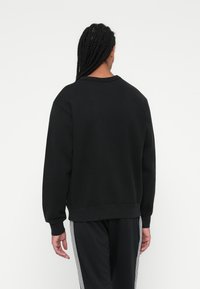 Sweatshirt preto com decote redondo e punhos canelados, combinado com calças pretas com detalhes em riscas brancas nas laterais.