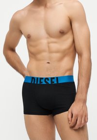 Homme torse nu avec un torse musclé portant un boxer noir avec une ceinture bleue marquée "DIESEL" sur fond blanc.