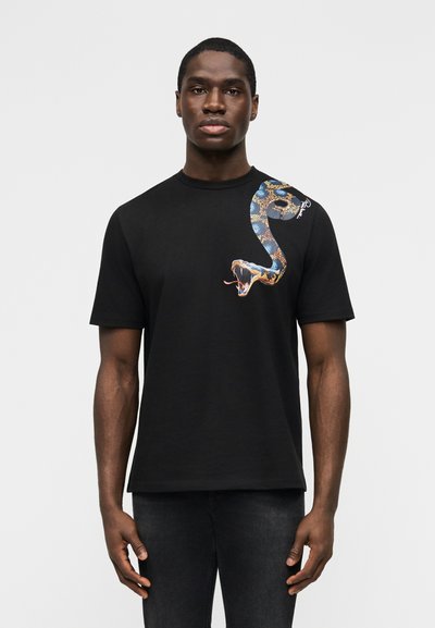 Jeune homme portant un t-shirt noir avec un motif détaillé et coloré de serpent s'enroulant de l'épaule à la poitrine.