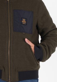 Giacca verde oliva con una tasca sul petto blu navy, zip dorata, polsini a costine e un logo ricamato sulla tasca. Tessuto testurizzato.