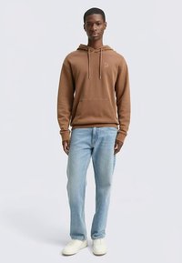 Sudadera marrón de algodón con capucha y bolsillo frontal, combinada con vaqueros azul claro y zapatillas blancas. La sudadera tiene un cordón ajustable y un detalle sutil del logo.