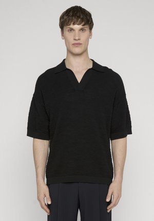 Młody mężczyzna o krótkich brązowych włosach, ubrany w czarną, teksturowaną koszulkę polo z krótkim rękawem i ciemne spodnie, stojący na tle jednolitego jasnego tła.