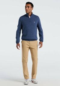 Man in een blauw half-zip sweater, beige broek en witte sneakers, staand met de armen iets van het lichaam af tegen een effen achtergrond.