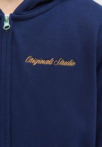 Marinblå zip-up hoodie med ribbad textur, med guldbroderad text "Originals Studio" på vänster bröst.