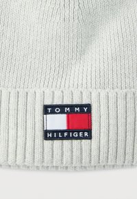 Lys gråstrikket lue med ribbet tekstur, med en rektangulær brodert lapp med ordene "TOMMY HILFIGER" og et fargeblokkdesign.