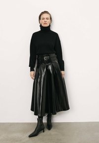 Suéter negro de cuello alto combinado con una falda de cuero negro plisada de cintura alta. Presenta botas de tobillo y un cinturón con un detalle de hebilla.