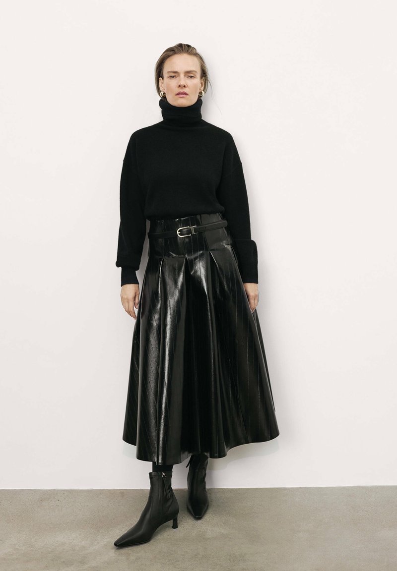 Crna pletena turtleneck majica, uparena s crnom visokoproširenom suknjom od patentnog materijala s nabranim detaljima, nošena sa čizmama s visokom potpeticom i uskim vrhom.