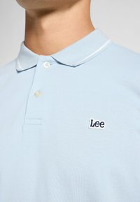 Ljusblå pikétröja av texturerat tyg. Har en klassisk krage, två knappar och en liten broderad "Lee"-logotyp i marinblått.