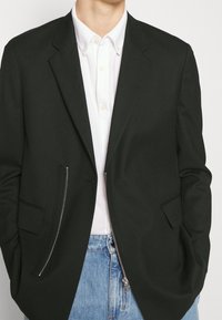 Schwarzer Blazer mit strukturiertem Design, ausgestattet mit Reißverschluss- und aufgesetzten Taschen, über einem weißen Hemd mit Kragen getragen. Sichtbare lässige Jeans.
