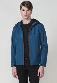 Giacca softshell blu con cappuccio, zip frontale, diverse tasche con zip, accenti arancioni a contrasto e polsini regolabili. Texture liscia.