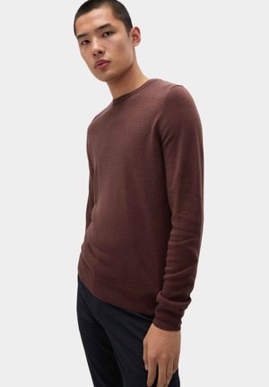 Pull en tricot marron avec un col rond et des manches longues, associé à un pantalon sombre. Le pull présente une texture lisse et une coupe ajustée.
