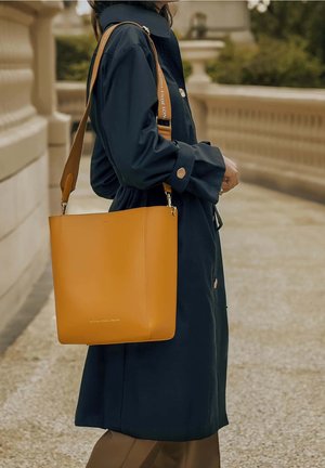 Gele leren totebag met een gladde afwerking, voorzien van een afneembare riem en zilverkleurige hardware, vastgehouden tegen een donkere jas als achtergrond.