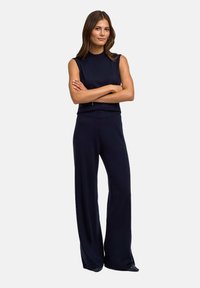 Navyblauwe mouwloze jumpsuit met hoge hals, brede pijpen en zachte textuur. Heeft een minimalistisch ontwerp en elegante lijnen.