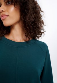 Teal sweatshirt met een geribde halslijn, een gladde textuur en raglanmouwen. Close-up weergave benadrukt de zachtheid van de stof en de kleurdetails.