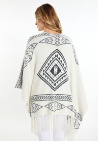 Poncho lavorato a maglia color crema con motivi geometrici neri, caratterizzato da un colletto scialle e un orlo con frange. Tessuto morbido e testurizzato.