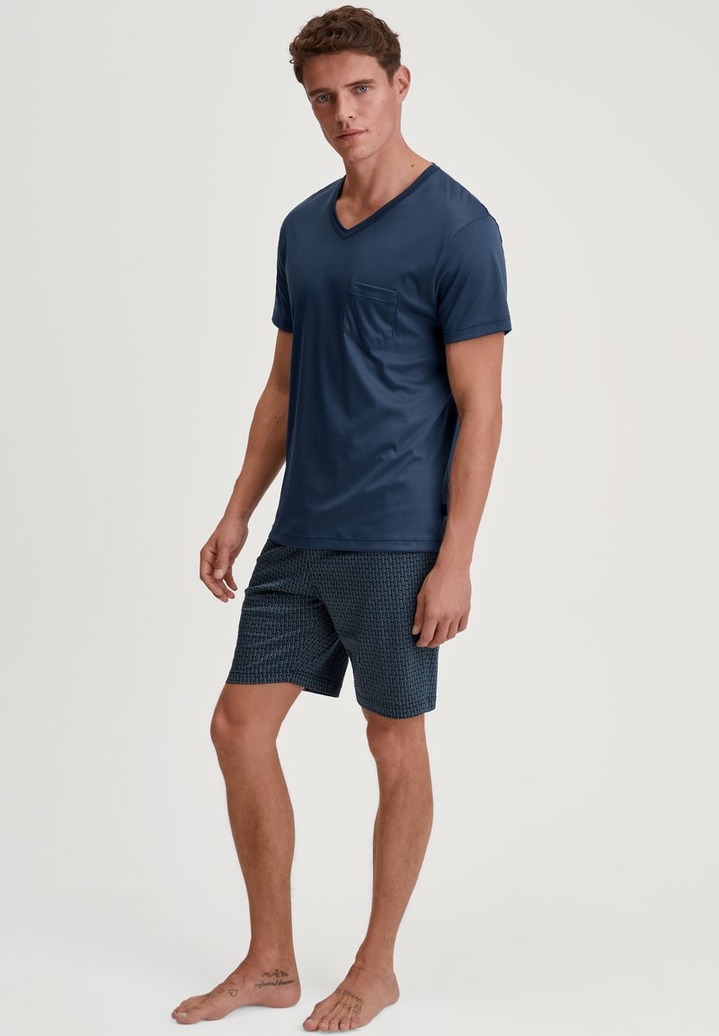 Navyblauw T-shirt met korte mouwen en V-hals met een borstzak, gecombineerd met gestreepte shorts in een vergelijkbare kleurenschema, op blote voeten gedragen.