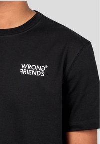 Černé bavlněné tričko s bílým nápisem "WRONG FRIENDS" na levé části hrudi, krátké rukávy a klasický kulatý výstřih.