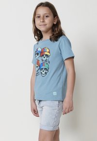 Camiseta de algodón azul con un gráfico de calaveras de dibujos animados con gorras coloridas. Combinada con pantalones cortos de mezclilla claros. Textura suave, mangas cortas.